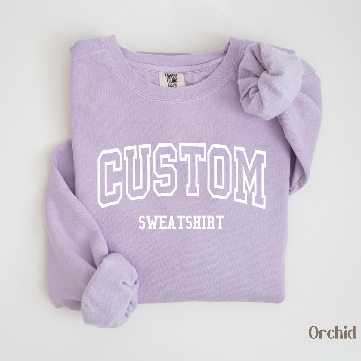 May include: A light purple sweatshirt with the word "CUSTOM" in white block letters above "SWEATSHIRT". The sleeves are folded, and the bottom right corner has the word "Orchid". The sweatshirt is a comfortable, casual style.