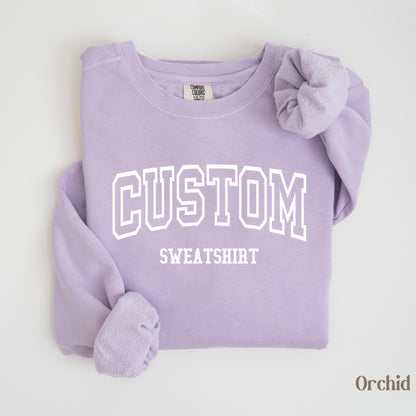 May include: A light purple sweatshirt with the word "CUSTOM" in white block letters above "SWEATSHIRT". The sleeves are folded, and the bottom right corner has the word "Orchid". The sweatshirt is a comfortable, casual style.