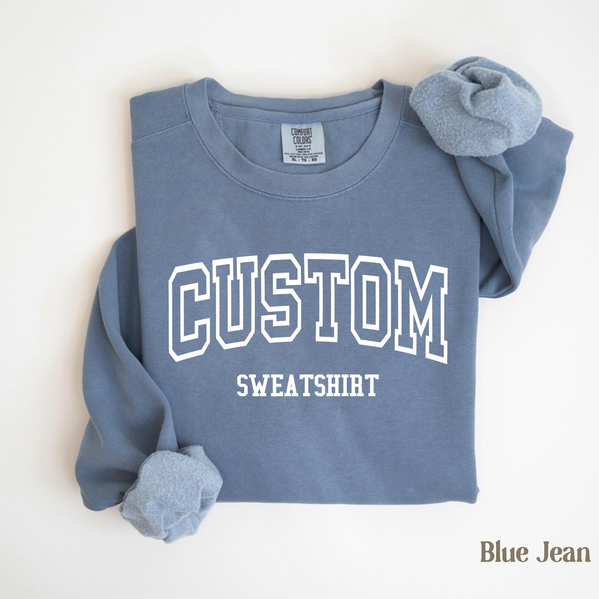 May include: A blue jean-colored sweatshirt with the word "CUSTOM" in white block letters above the word "SWEATSHIRT". The sweatshirt is folded, with the sleeves and collar visible. The Comfort Colors tag is visible.