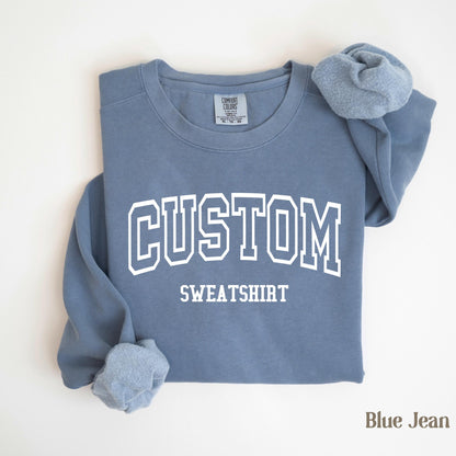 May include: A blue jean-colored sweatshirt with the word "CUSTOM" in white block letters above the word "SWEATSHIRT". The sweatshirt is folded, with the sleeves and collar visible. The Comfort Colors tag is visible.
