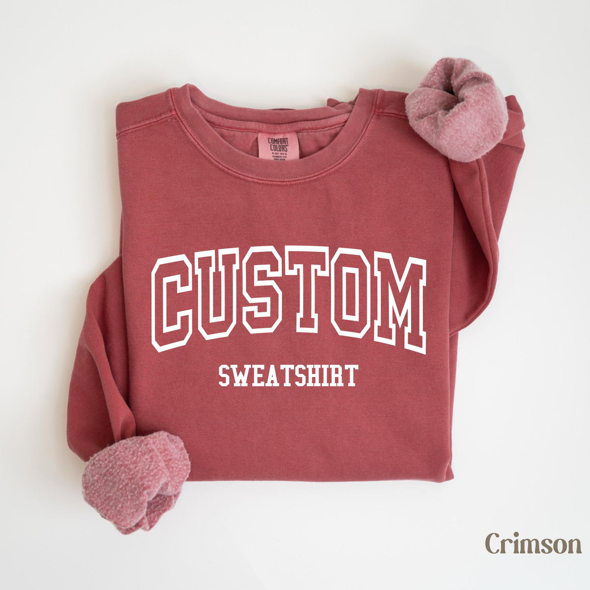 May include: Crimson-colored sweatshirt with the word "CUSTOM" in white block letters above "SWEATSHIRT". The sweatshirt has a crew neck and ribbed cuffs. The sleeves are folded, revealing a soft, pinkish lining. The word "Crimson" is in the bottom right corner.