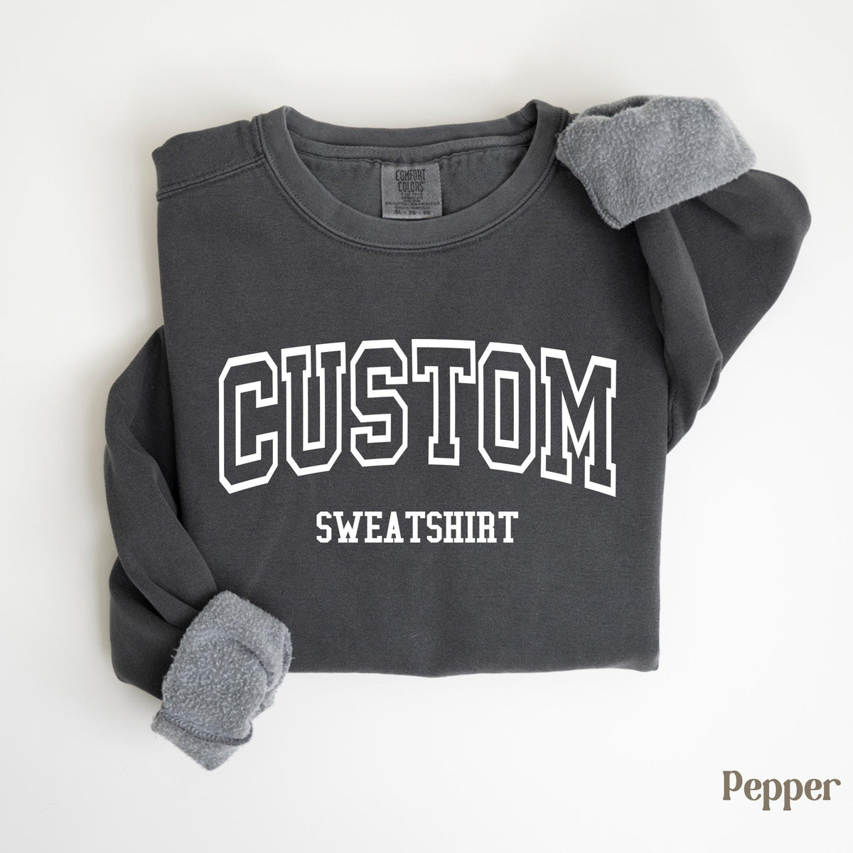 May include: A dark gray sweatshirt with the word "CUSTOM" in large white block letters above the word "SWEATSHIRT". The sleeves are cuffed with a soft, gray material. The word "Pepper" is in the bottom right corner.