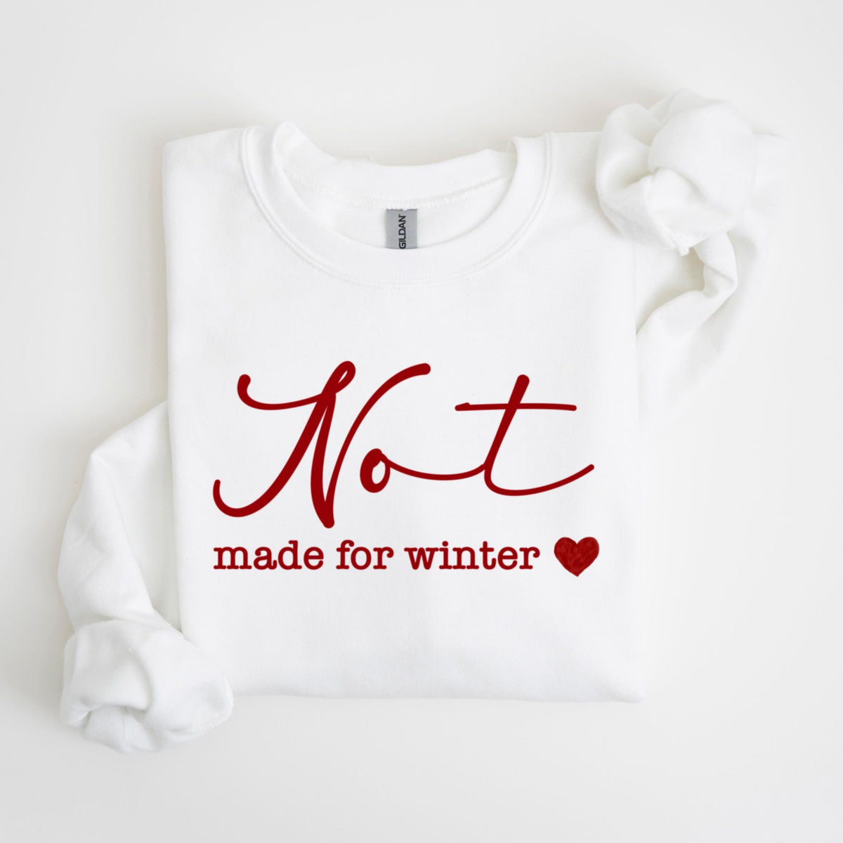 May include: White sweatshirt with the red text "Not made for winter" and a red heart. The sweatshirt is folded, with the sleeves visible. The Gildan brand tag is visible on the collar.