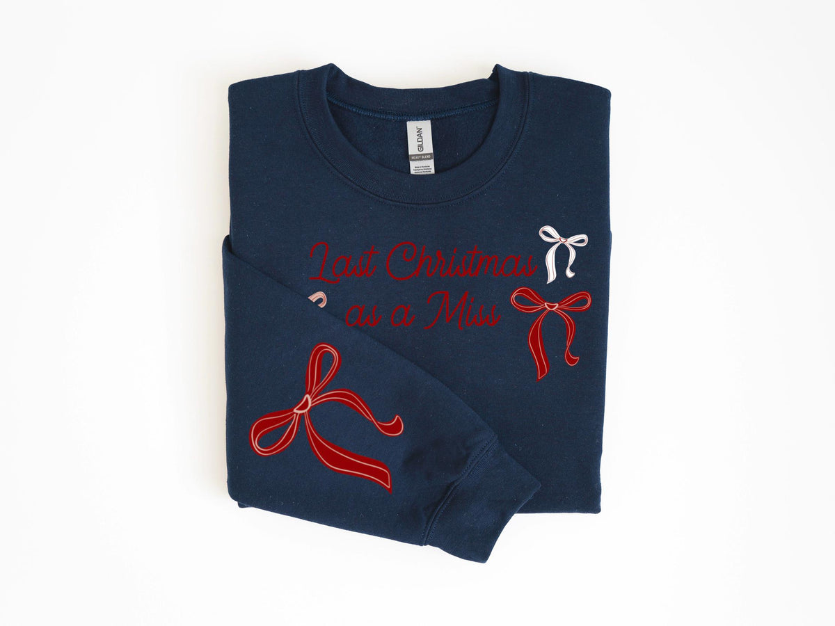 May include: Navy blue sweatshirt with the text "Last Christmas as a Miss" in red script. The sweatshirt features red and white bow designs. A cozy, festive garment for the holiday season.