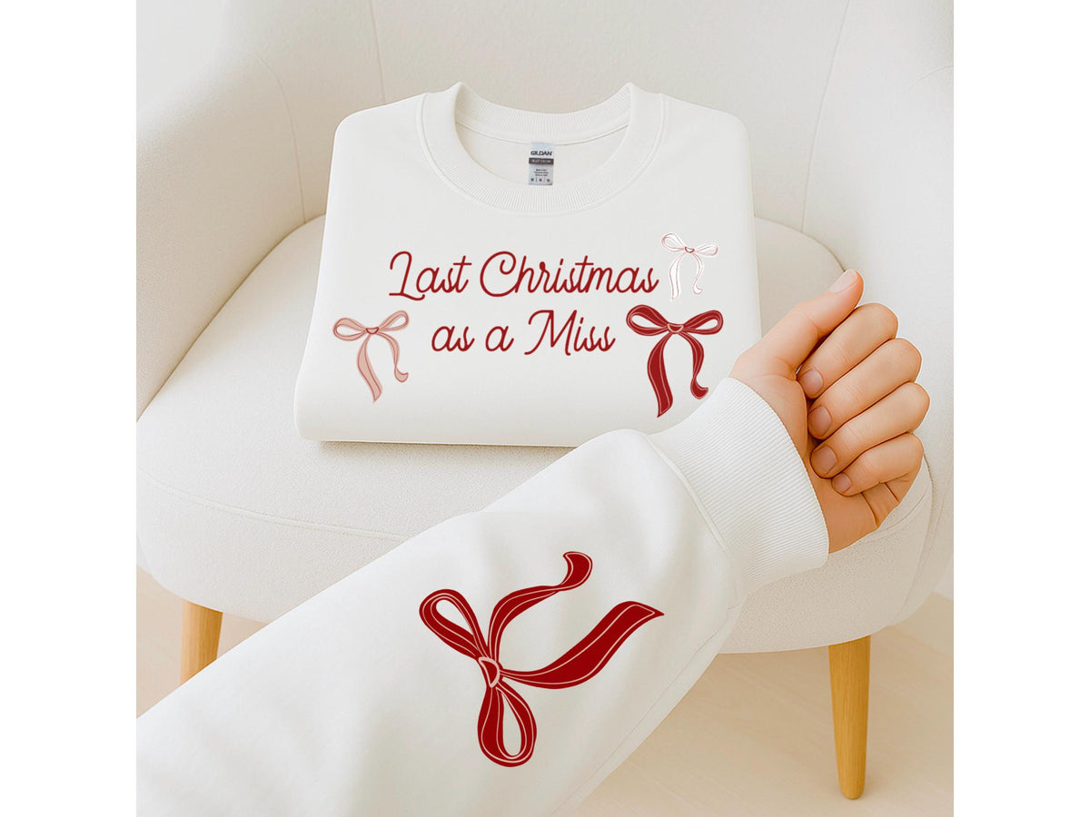 May include: A white sweatshirt with the text "Last Christmas as a Miss" in burgundy script, accented with bow graphics. A sleeve of a matching sweatshirt is visible, featuring a large burgundy bow design. The sweatshirt is folded on a white chair.