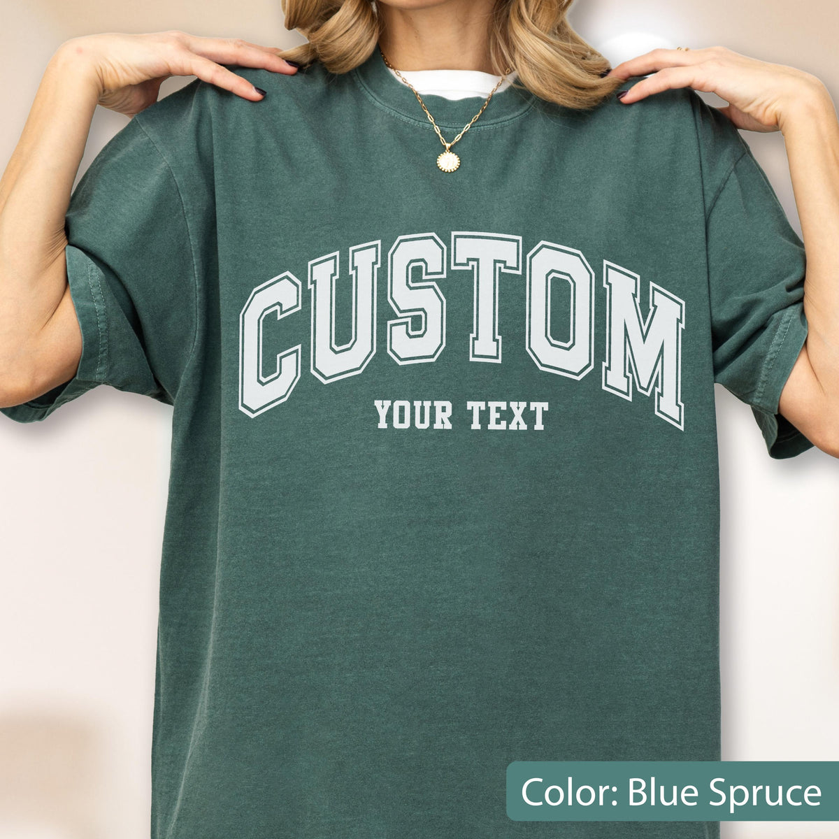 May include: Blue spruce-colored t-shirt with the word "CUSTOM" in white block letters arched across the chest. Below the word "CUSTOM" is the text "YOUR TEXT" in a smaller font. The shirt has short sleeves and a crew neck.