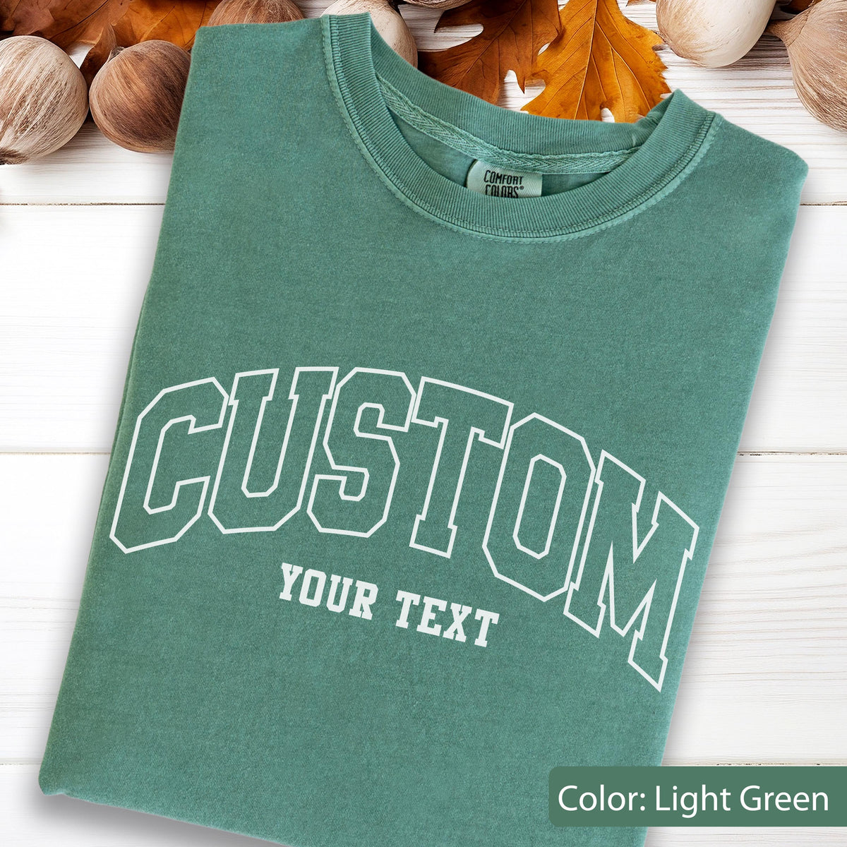 May include: Light green t-shirt with the word "CUSTOM" arched across the front in white block letters. Below the word "CUSTOM" is the text "YOUR TEXT" in white block letters. The shirt is folded and has a crew neck.
