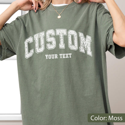 May include: A moss green t-shirt with the word "CUSTOM" arched across the chest in white, block letters. Below the word "CUSTOM" is the text "YOUR TEXT" in a smaller font. The shirt is a solid color and has short sleeves.