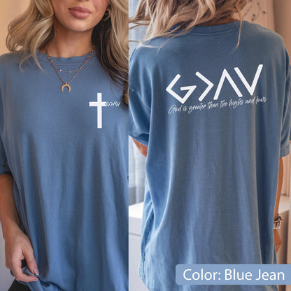 May include: A blue jean colored t-shirt featuring a white cross graphic on the front left chest. The back of the shirt displays the white text G > ^ v with the phrase God is greater than the highs and lows printed underneath in white script.