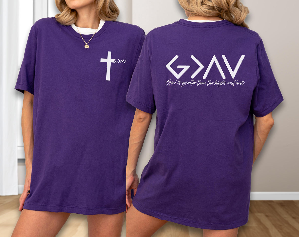 May include: Purple t-shirt with a white cross and the letters "G>AV" on the front. The back of the shirt features the text "G>AV" and the phrase "God is greater than the highs and lows" in white. The shirt has a white collar.
