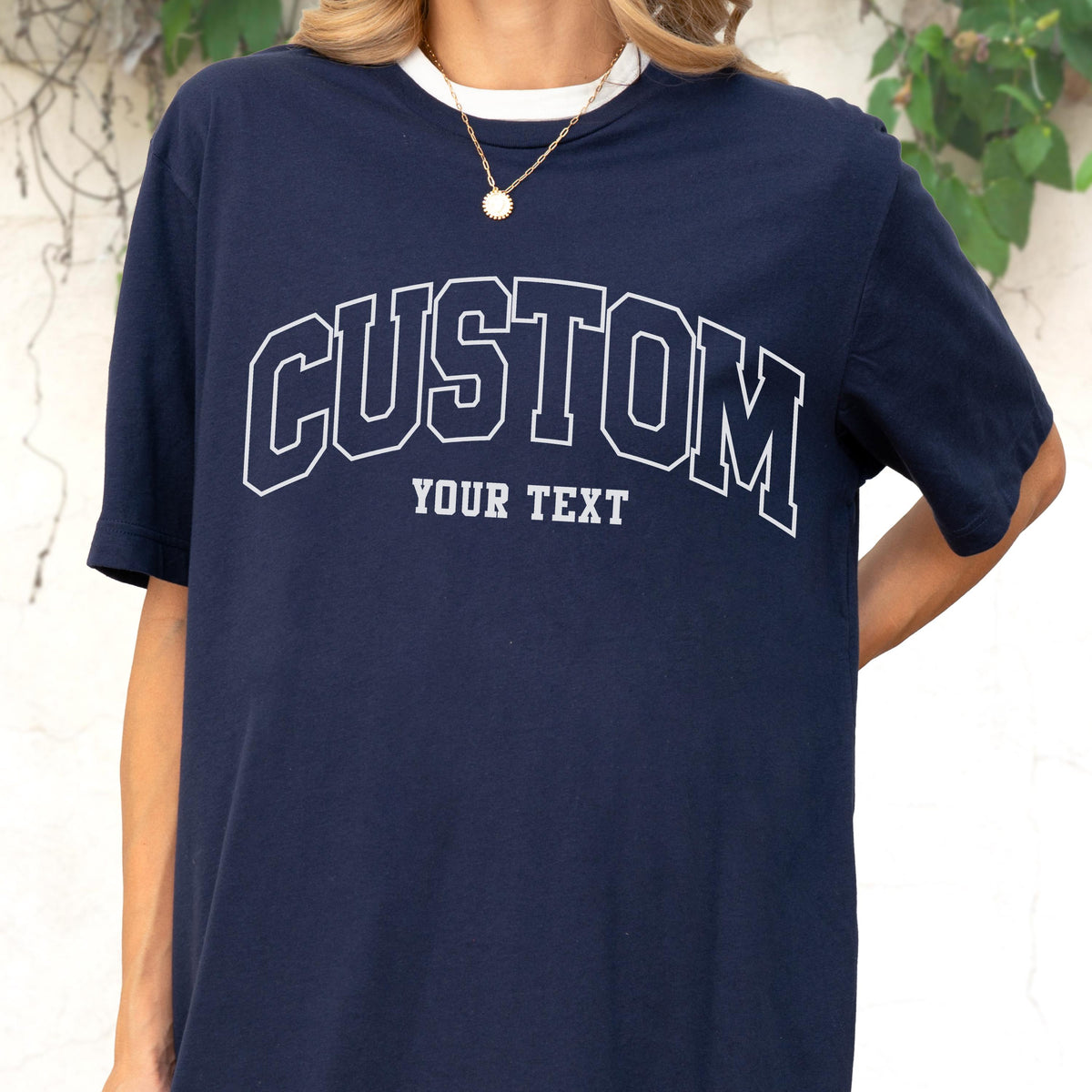May include: Navy blue t-shirt with a white collar and the word "CUSTOM" arched across the chest in a collegiate font. Below the word "CUSTOM" is the text "YOUR TEXT". The shirt is a short-sleeved design.