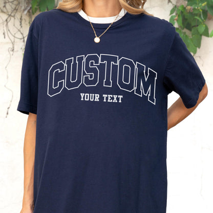 May include: Navy blue t-shirt with a white collar and the word "CUSTOM" arched across the chest in a collegiate font. Below the word "CUSTOM" is the text "YOUR TEXT". The shirt is a short-sleeved design.