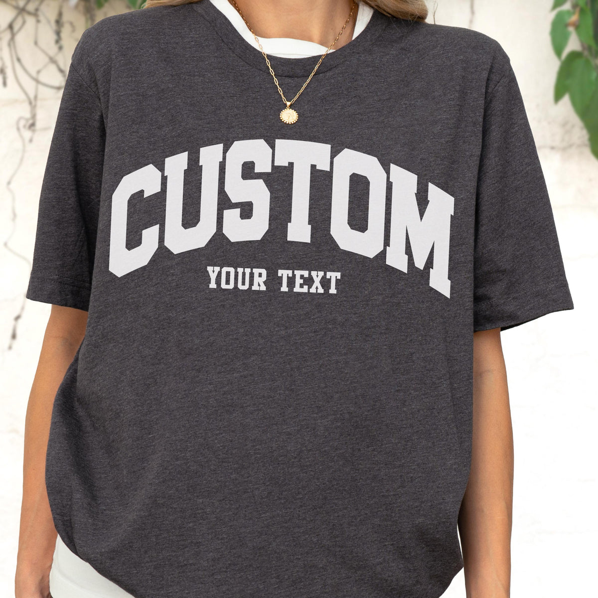 May include: Dark gray t-shirt with white text that reads "CUSTOM" above "YOUR TEXT" in a collegiate font. The shirt has a white collar and short sleeves. A gold necklace is visible.