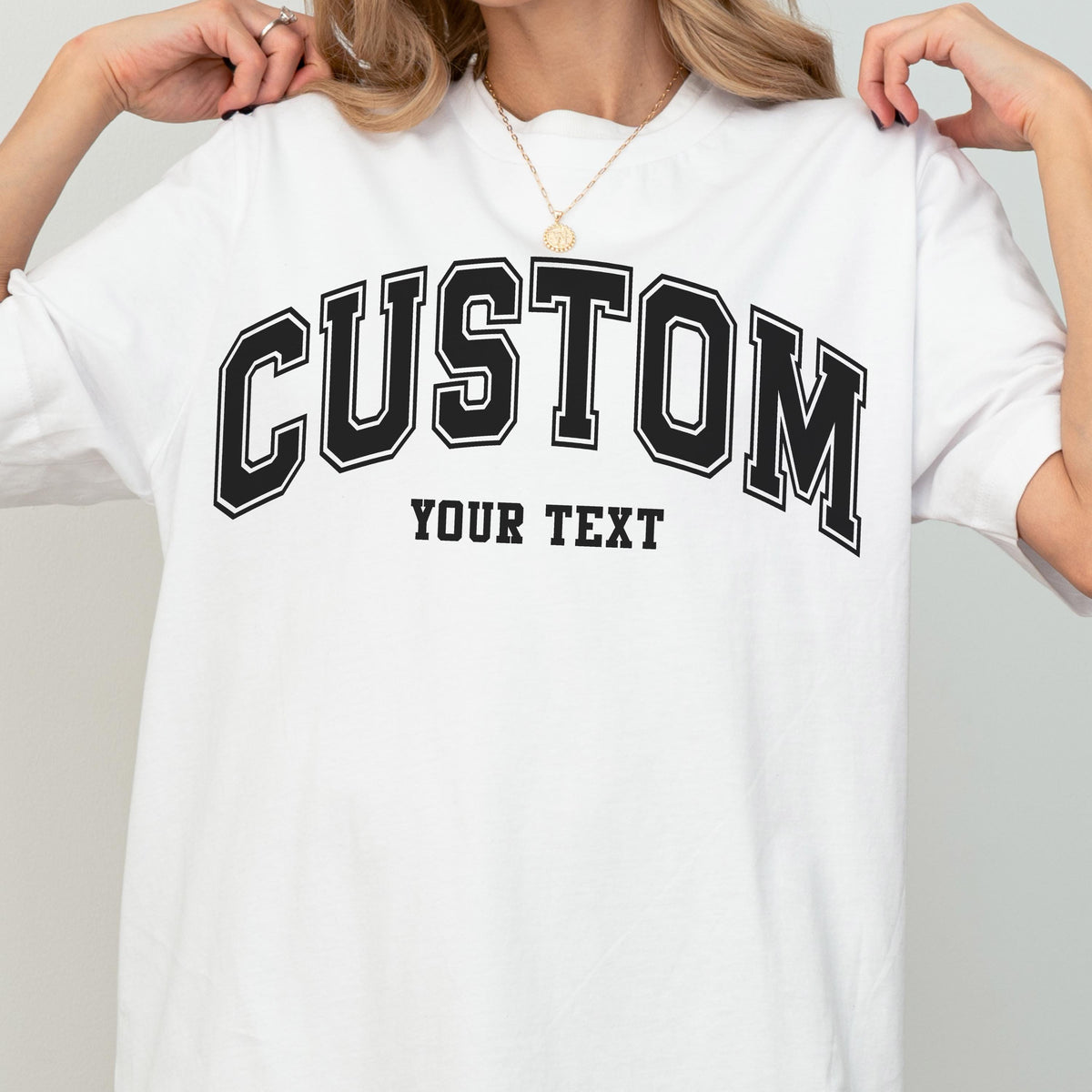May include: White t-shirt with the word "CUSTOM" in large, black, collegiate-style lettering across the chest. Below the word "CUSTOM" is the text "YOUR TEXT" in a smaller, block font. The shirt has a crew neck and short sleeves.