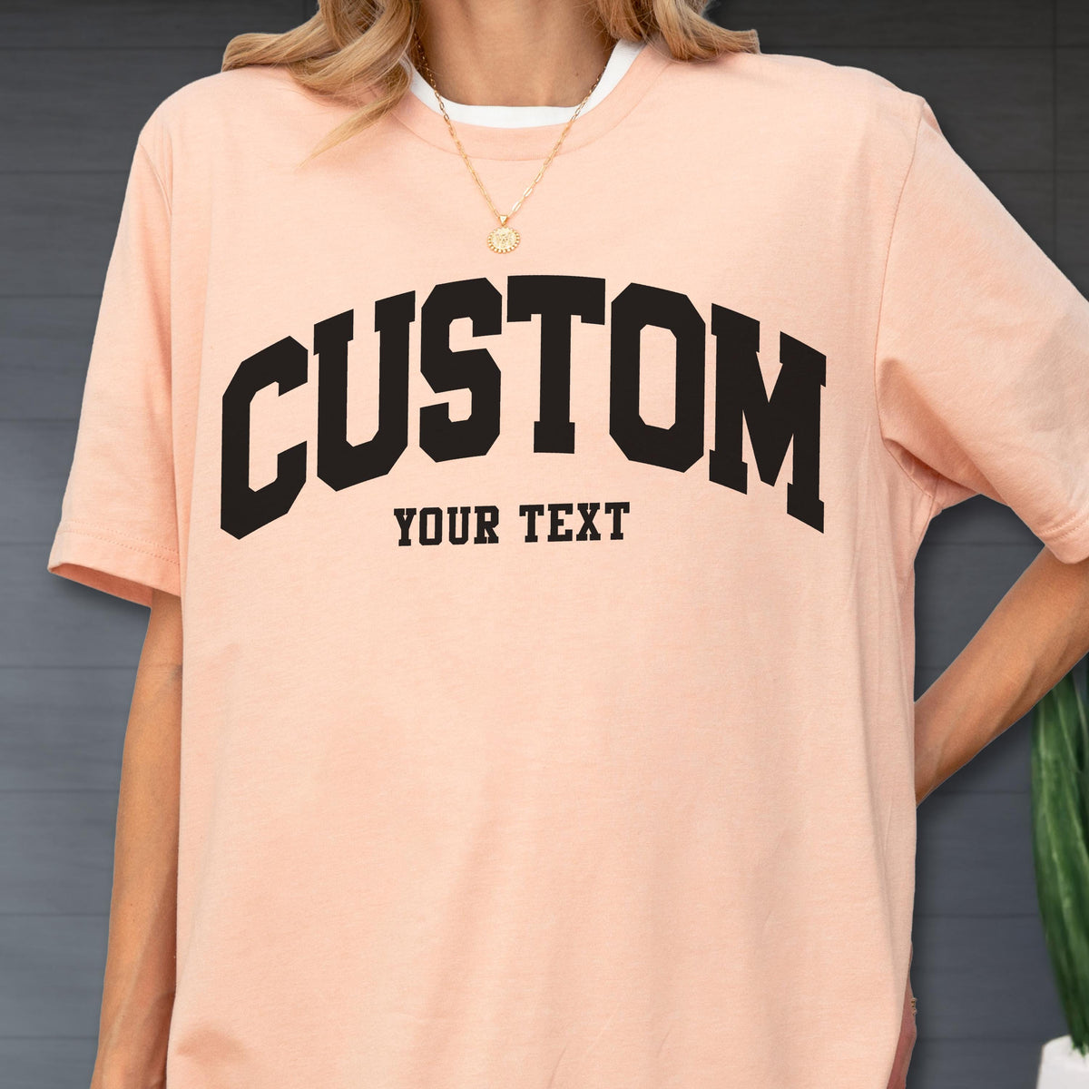 May include: A light peach-colored t-shirt with the word "CUSTOM" in bold, black, arched lettering. Below, the text "YOUR TEXT" is printed in a smaller, blocky font. The shirt has a white crew neck collar.