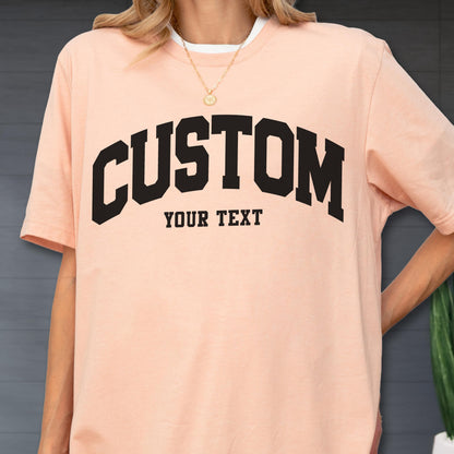 May include: A light peach-colored t-shirt with the word "CUSTOM" in bold, black, arched lettering. Below, the text "YOUR TEXT" is printed in a smaller, blocky font. The shirt has a white crew neck collar.