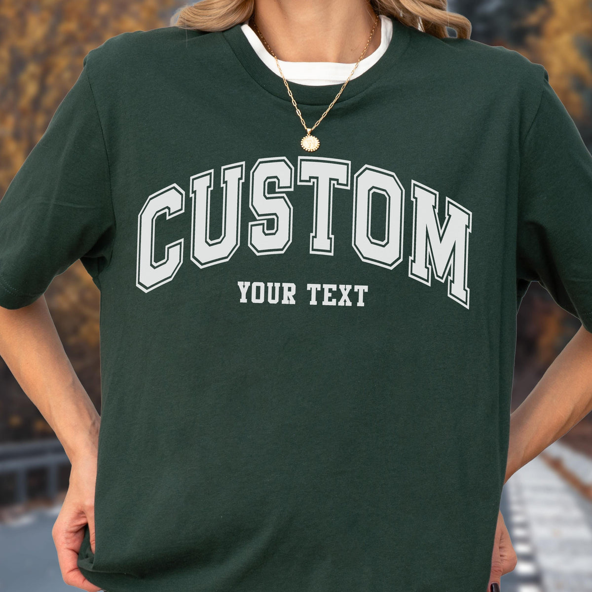May include: Dark green t-shirt with the word "CUSTOM" in white, arched across the chest. Below the word "CUSTOM" is the text "YOUR TEXT". The shirt has a white collar and is worn with a gold necklace.