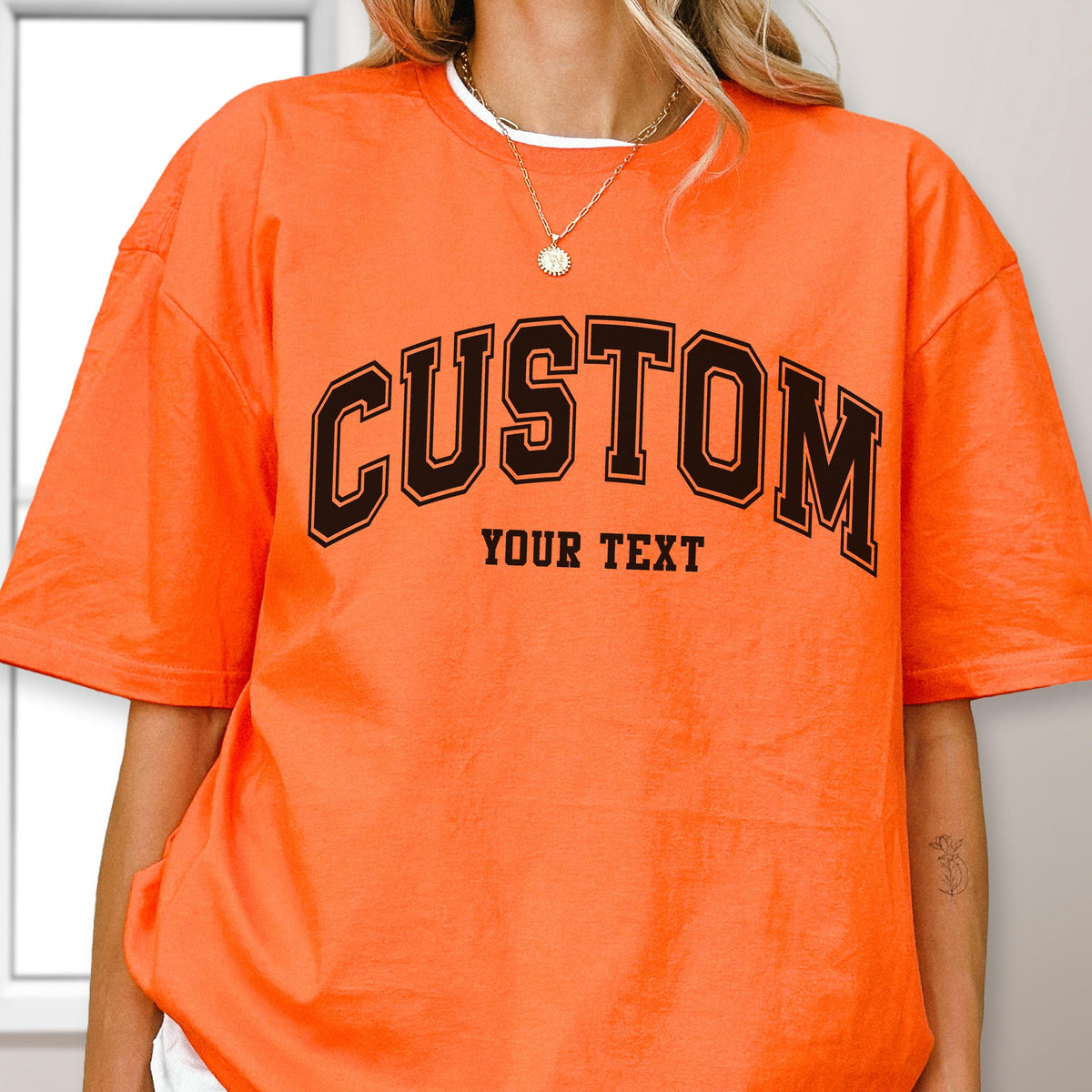 May include: Bright orange t-shirt with the word "CUSTOM" in black block letters with a black outline. Below the word "CUSTOM" is the text "YOUR TEXT". The shirt has short sleeves and a crew neck.