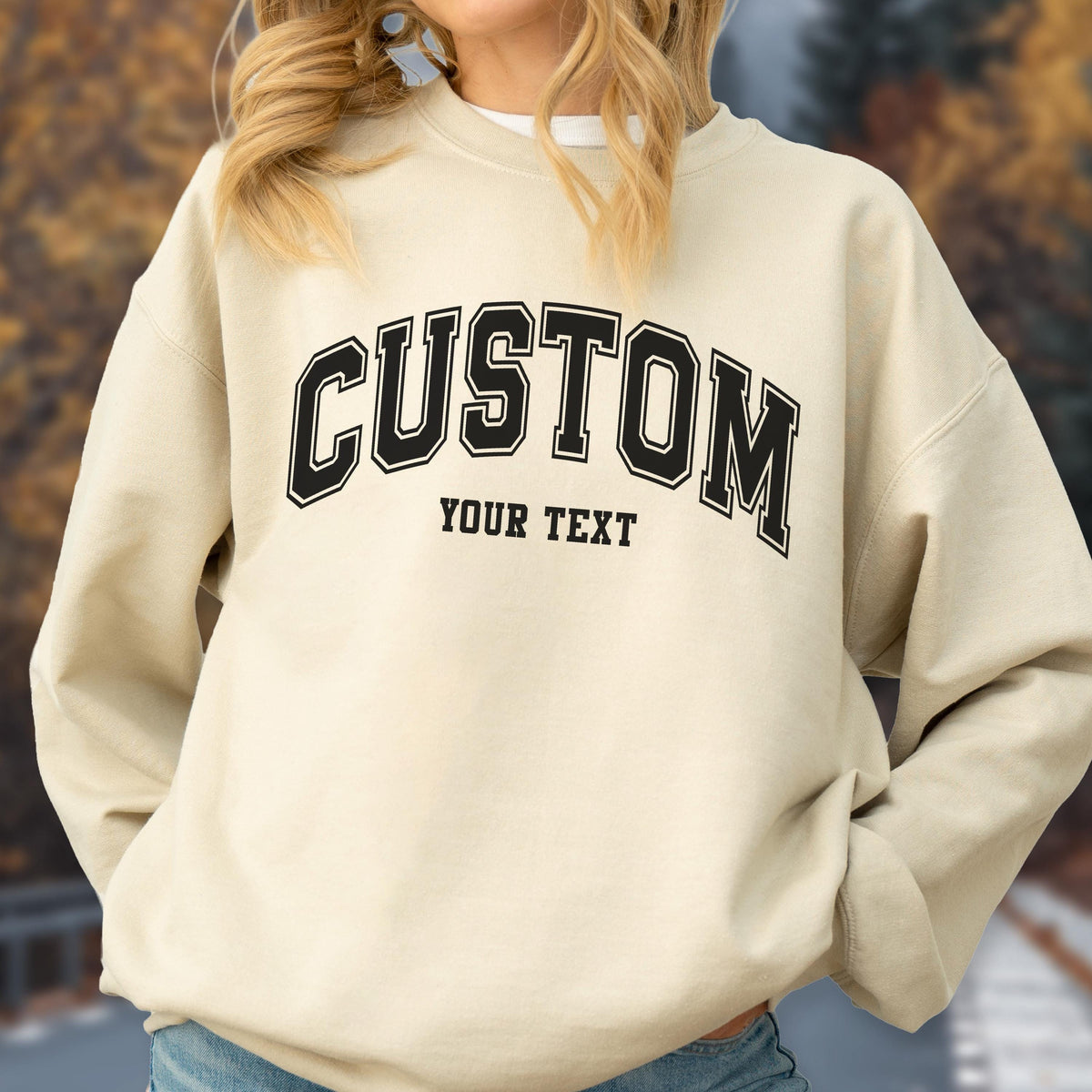 May include: A cream-colored sweatshirt with the word "CUSTOM" arched across the chest in black block letters with a white outline. Below the word "CUSTOM" is the text "YOUR TEXT" in a smaller, black font. The sweatshirt is a casual style.