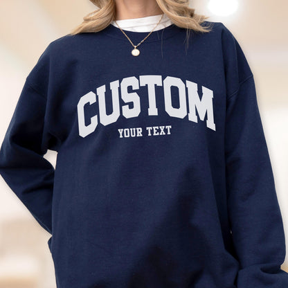 May include: Navy blue sweatshirt with the word "CUSTOM" in large, white, arched letters across the chest. Below the word "CUSTOM" is the text "YOUR TEXT" in a smaller, white font. The sweatshirt is a solid color.