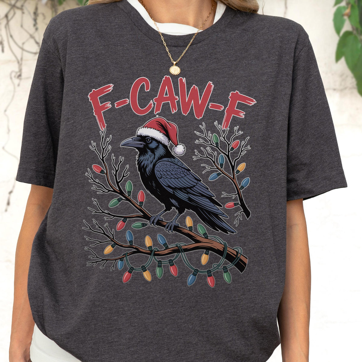 May include: A dark heather gray t-shirt featuring a graphic of a black crow wearing a red and white Santa hat, perched on a brown tree branch wrapped in colorful Christmas lights. The text F-CAW-F is printed in red above the crow illustration.
