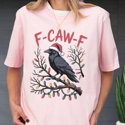 May include: A light pink t-shirt featuring a graphic of a black crow wearing a red and white Santa hat, perched on a bare tree branch wrapped in colorful Christmas lights, with the red text F-CAW-F printed in a distressed font above the bird.