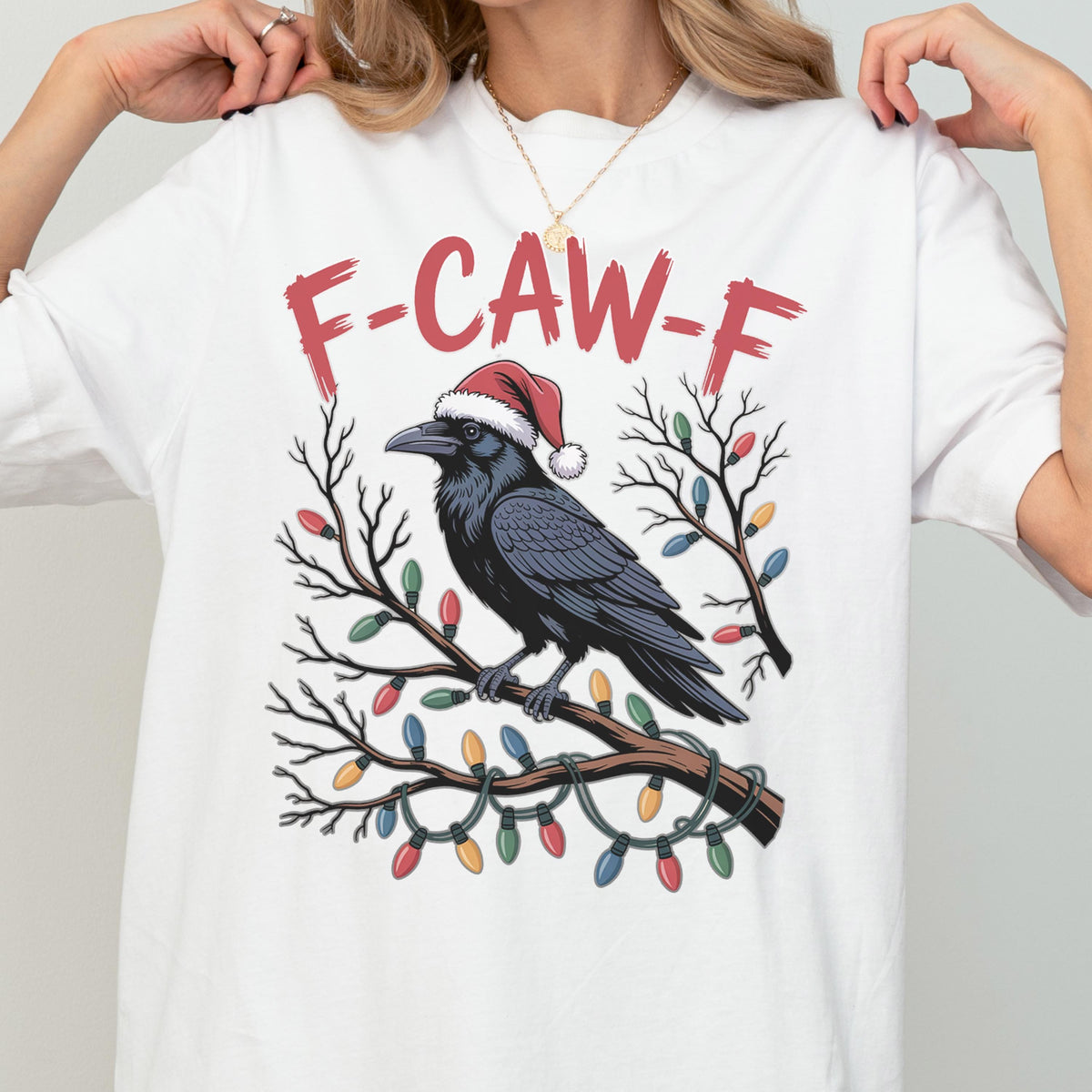 May include: A white t-shirt featuring a graphic of a black crow wearing a red and white Santa hat, perched on a bare tree branch wrapped in colorful Christmas lights. The text F-CAW-F is printed in red above the crow in a distressed font.