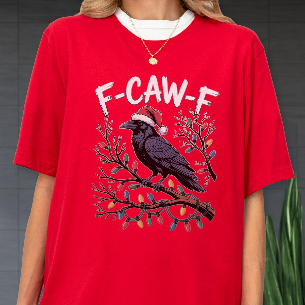 May include: A bright red t-shirt featuring a graphic of a black raven wearing a red and white Santa hat, perched on a branch wrapped in colorful Christmas lights. The text F-CAW-F is printed in white above the raven on the front of the shirt.