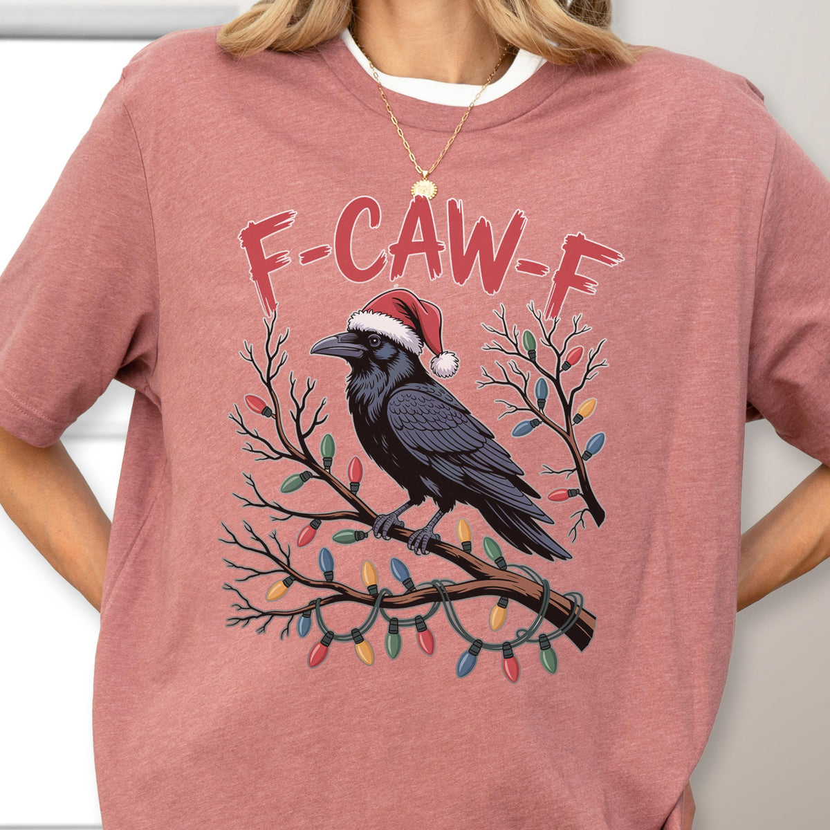 May include: A dusty rose short-sleeve t-shirt featuring a graphic of a black crow wearing a red and white Santa hat, perched on a bare tree branch wrapped in colorful Christmas lights, with the text F-CAW-F printed in red above the crow.