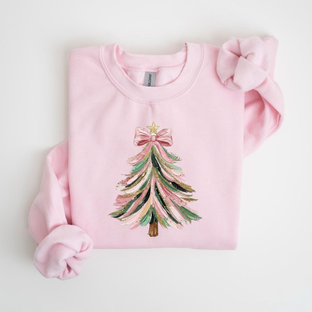 May include: A light pink sweatshirt featuring a Christmas tree design. The tree is decorated with pink, green, gold, and black brushstrokes, topped with a pink bow and a gold star. The sweatshirt has a crew neck and long sleeves.
