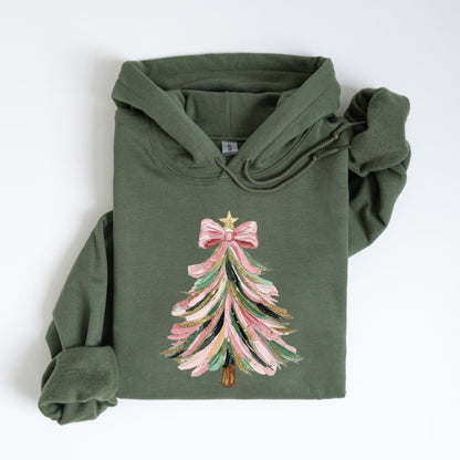 May include: Olive green hoodie featuring a Christmas tree design. The tree is decorated with pink, green, and gold brushstrokes, topped with a pink bow and a gold star. The hoodie has a drawstring hood and long sleeves.