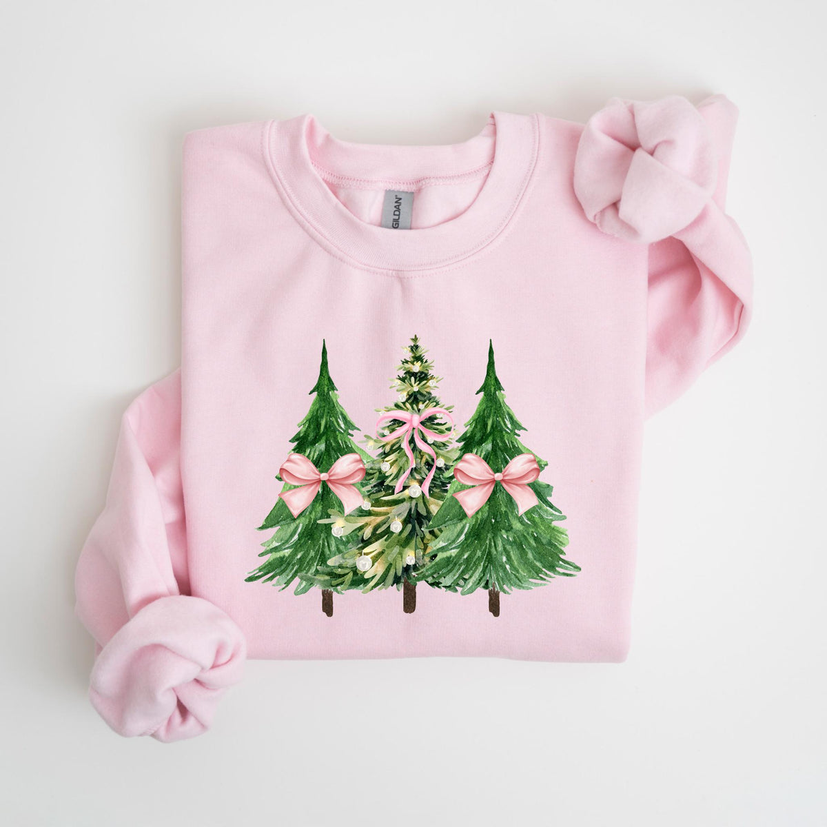 May include: A light pink sweatshirt featuring a watercolor design of three Christmas trees. The trees are green with pink bows, and the middle tree has a more detailed design. The sweatshirt is made of a soft material.