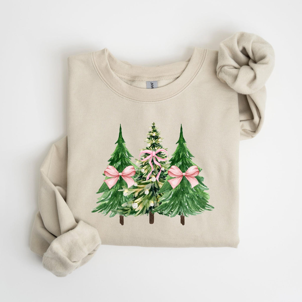 May include: A beige sweatshirt featuring a watercolor design of three Christmas trees. The trees are green with pink bows, and the middle tree has a pink ribbon. The sweatshirt has long sleeves and a matching scrunchie.