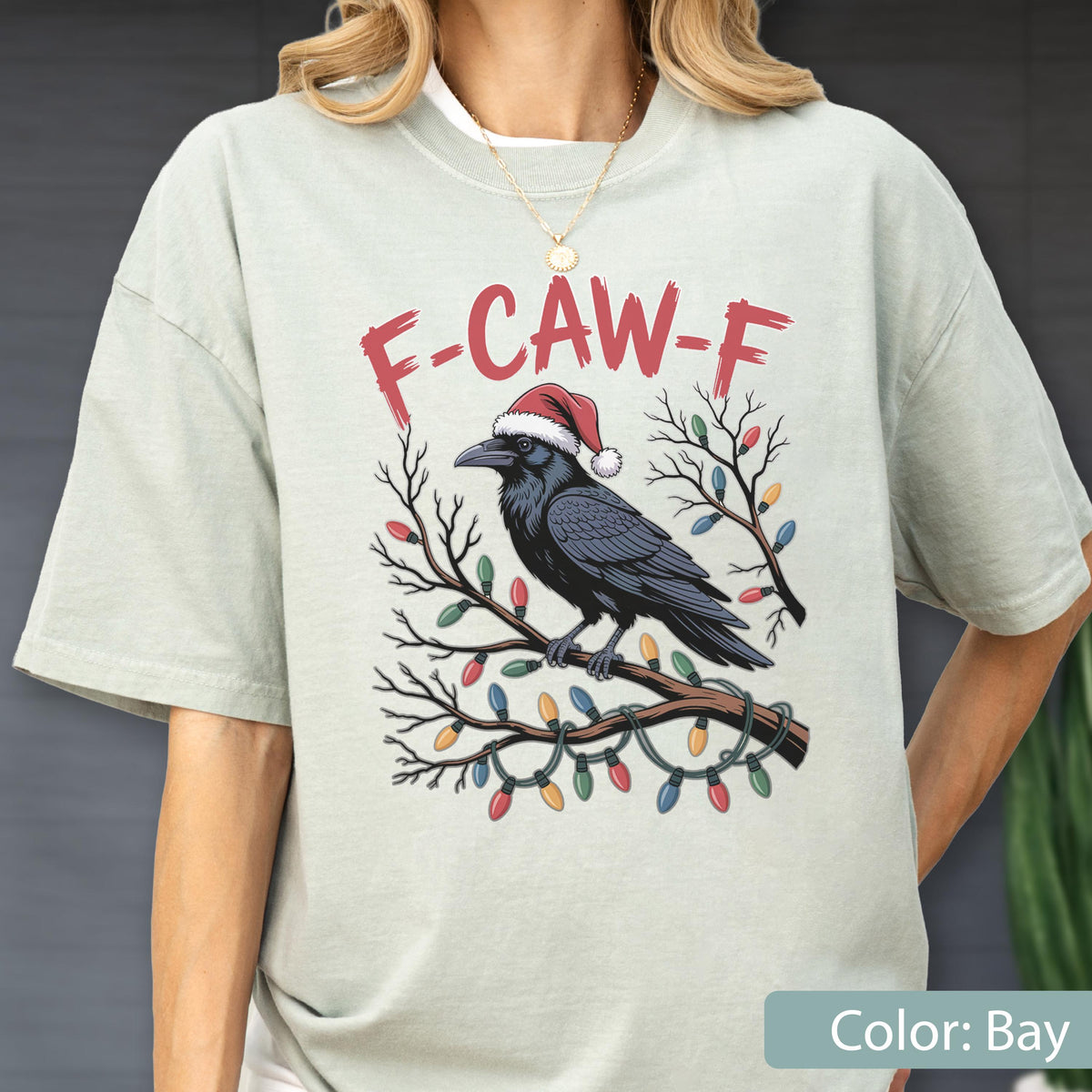 May include: A bay-colored t-shirt featuring a graphic of a black crow wearing a red and white Santa hat, perched on a branch wrapped in colorful Christmas lights. Above the crow, the text F-CAW-F is printed in a distressed red font.