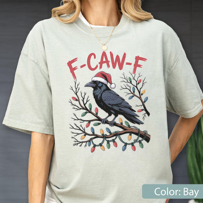 May include: A bay-colored t-shirt featuring a graphic of a black crow wearing a red and white Santa hat, perched on a branch wrapped in colorful Christmas lights. Above the crow, the text F-CAW-F is printed in a distressed red font.