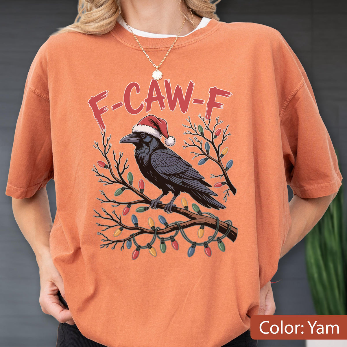 May include: A yam-colored t-shirt featuring a graphic of a black crow wearing a red and white Santa hat perched on a bare tree branch wrapped in colorful Christmas lights, with the red text F-CAW-F printed in an arc above the bird.