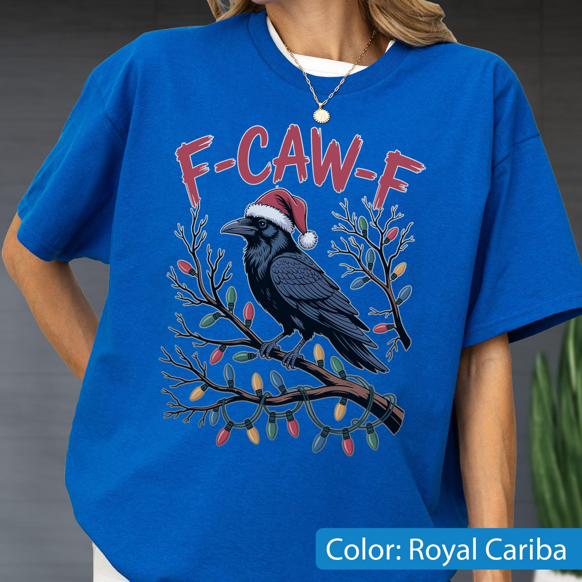 May include: A royal blue t-shirt featuring a graphic of a black raven wearing a red and white Santa hat, perched on a tree branch wrapped in colorful Christmas lights. The text F-CAW-F is printed in red above the raven. The shirt has a crew neckline.