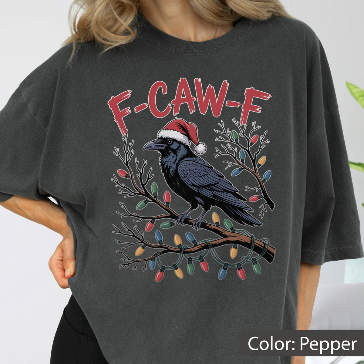 May include: A charcoal gray t-shirt featuring a graphic of a black crow wearing a red and white Santa hat, perched on a branch wrapped in colorful Christmas lights. Above the crow, the red text reads F-CAW-F in a distressed, jagged font style.