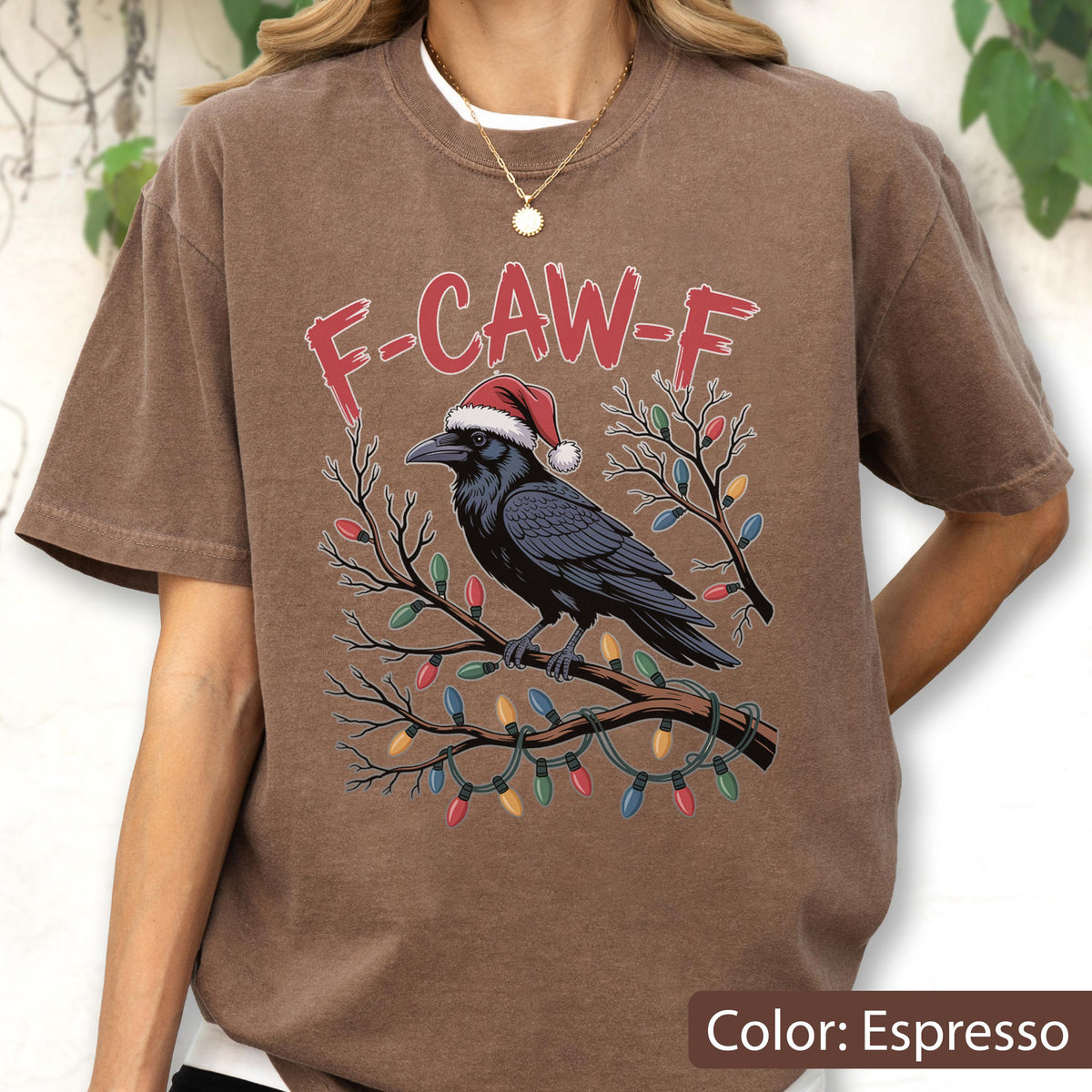 May include: An espresso-colored t-shirt featuring a graphic of a black crow wearing a red and white Santa hat, perched on a branch wrapped in colorful Christmas lights. The text F-CAW-F is printed in red above the crow in a distressed font.