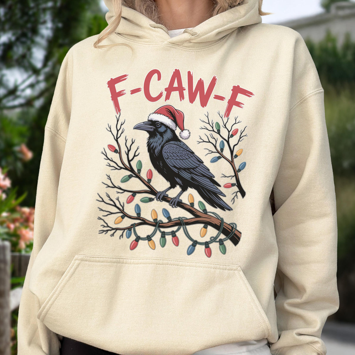 May include: A cream-colored hooded sweatshirt featuring a graphic of a black raven wearing a red and white Santa hat, perched on a branch wrapped in colorful Christmas lights, with the red text F-CAW-F printed above the bird illustration.