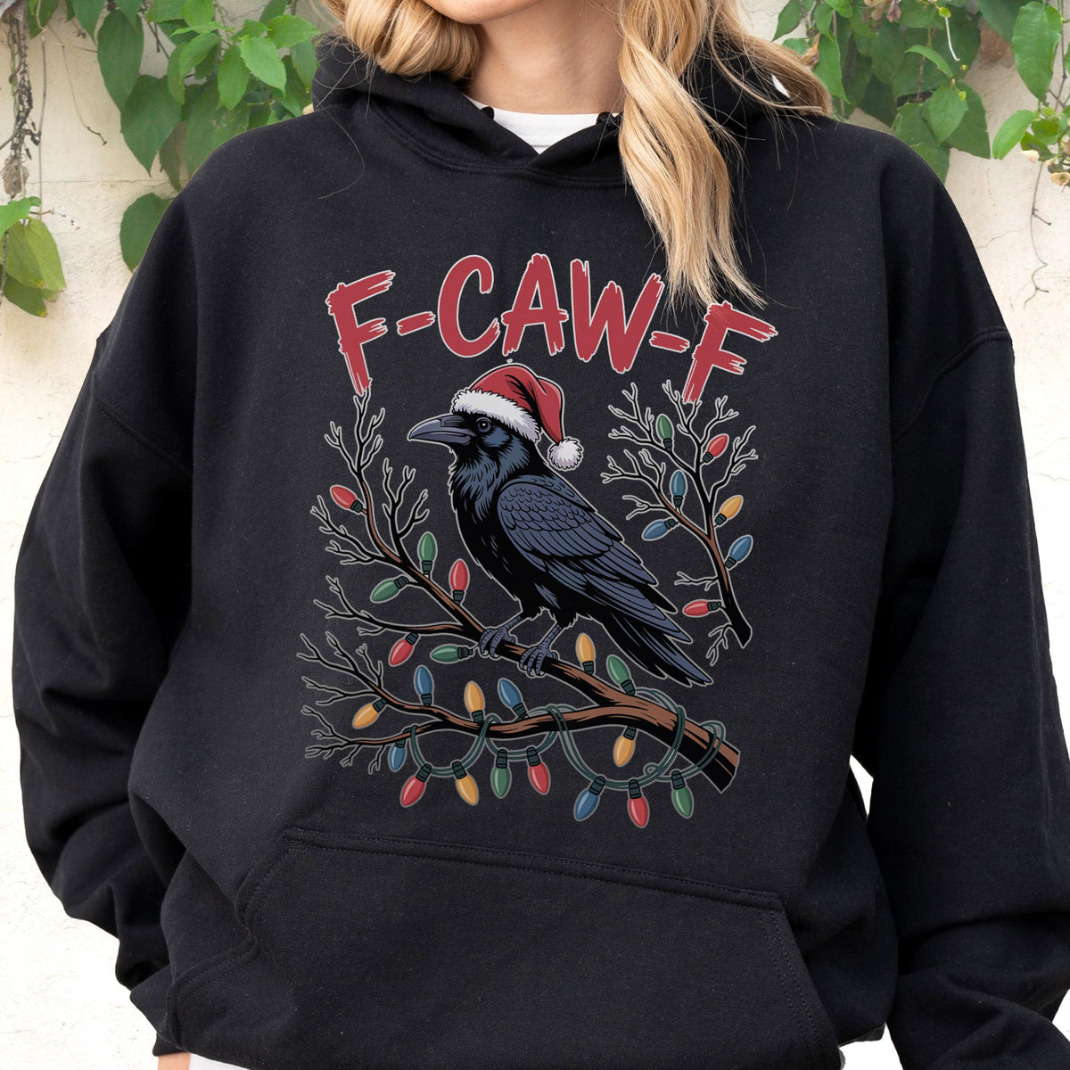 May include: A black hooded sweatshirt featuring a graphic of a raven wearing a red and white Santa hat, perched on a branch wrapped in colorful Christmas lights. The text F-CAW-F is printed in red above the raven, which has black feathers.