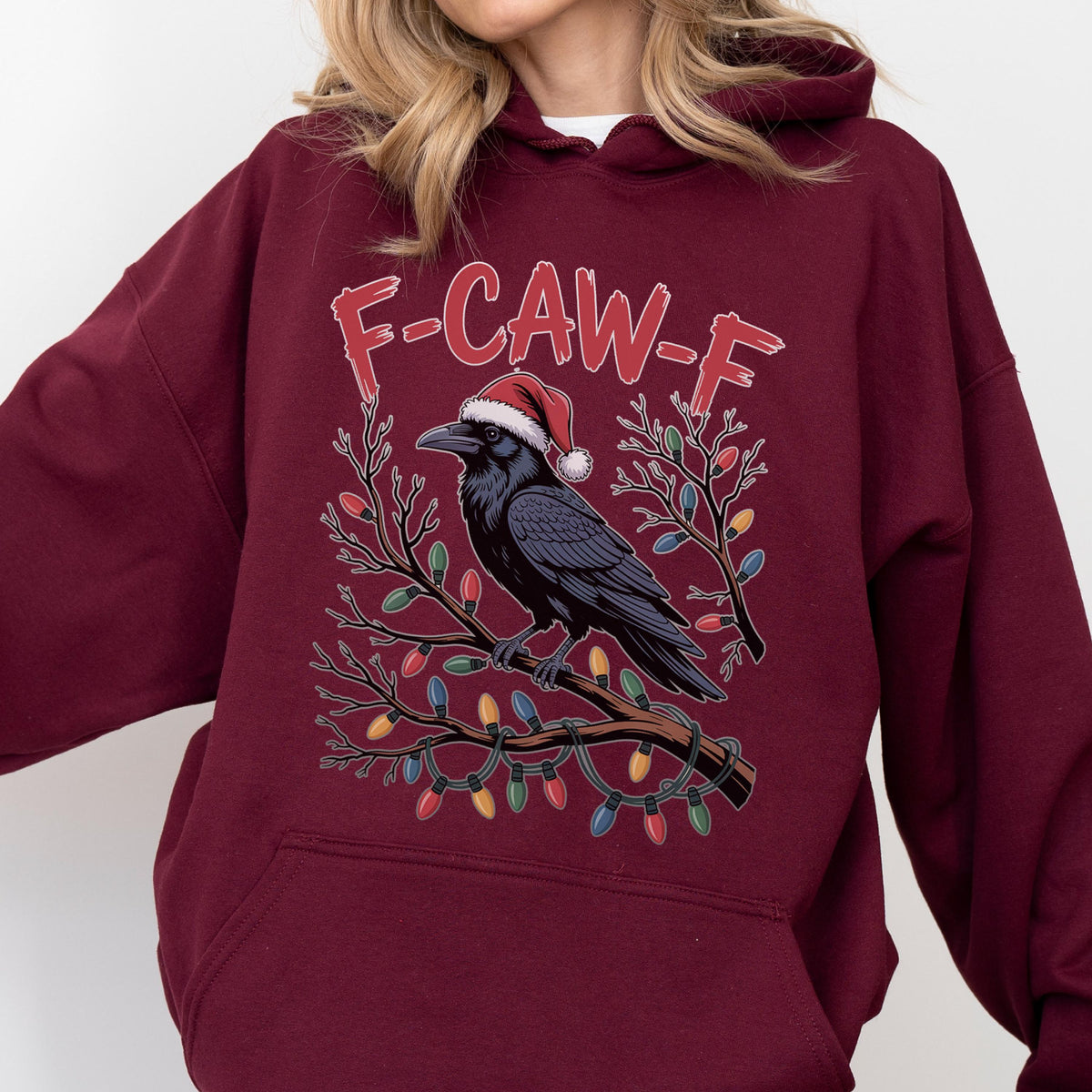 May include: A maroon hooded sweatshirt featuring a graphic of a black crow wearing a red and white Santa hat, perched on a tree branch wrapped in colorful Christmas lights. The text F-CAW-F is printed in red above the crow on the front of the garment.