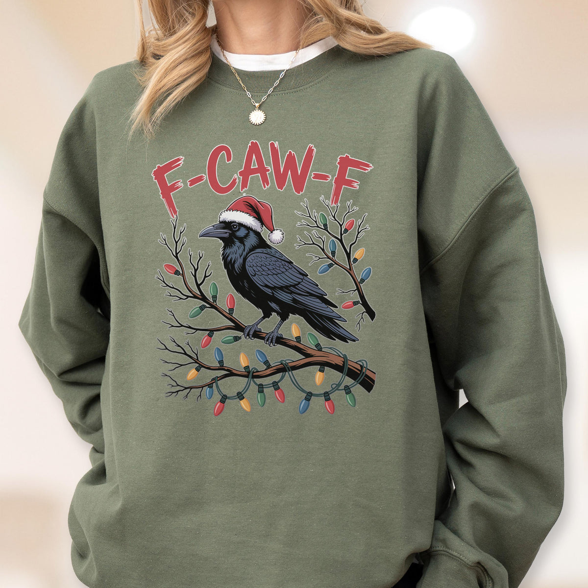May include: An olive green crewneck sweatshirt featuring a graphic of a black crow wearing a red and white Santa hat, perched on a branch wrapped in colorful Christmas lights. The text F-CAW-F is printed in red above the crow illustration.