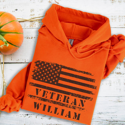 May include: Orange hoodie featuring a distressed American flag design with the text "VETERAN WILLIAM" in black. The hoodie has a drawstring hood and a front pocket. A pumpkin is visible in the upper left corner.