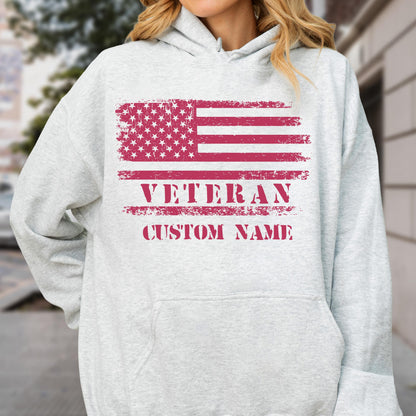 May include: A light gray hoodie with a front pocket and a drawstring hood. The design features a distressed American flag in red and white, with the words "VETERAN" and "CUSTOM NAME" printed below in red.