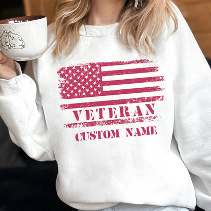 May include: White sweatshirt featuring a distressed American flag design in red. The word "VETERAN" is printed below the flag, with "CUSTOM NAME" underneath. The sweatshirt is a casual style.