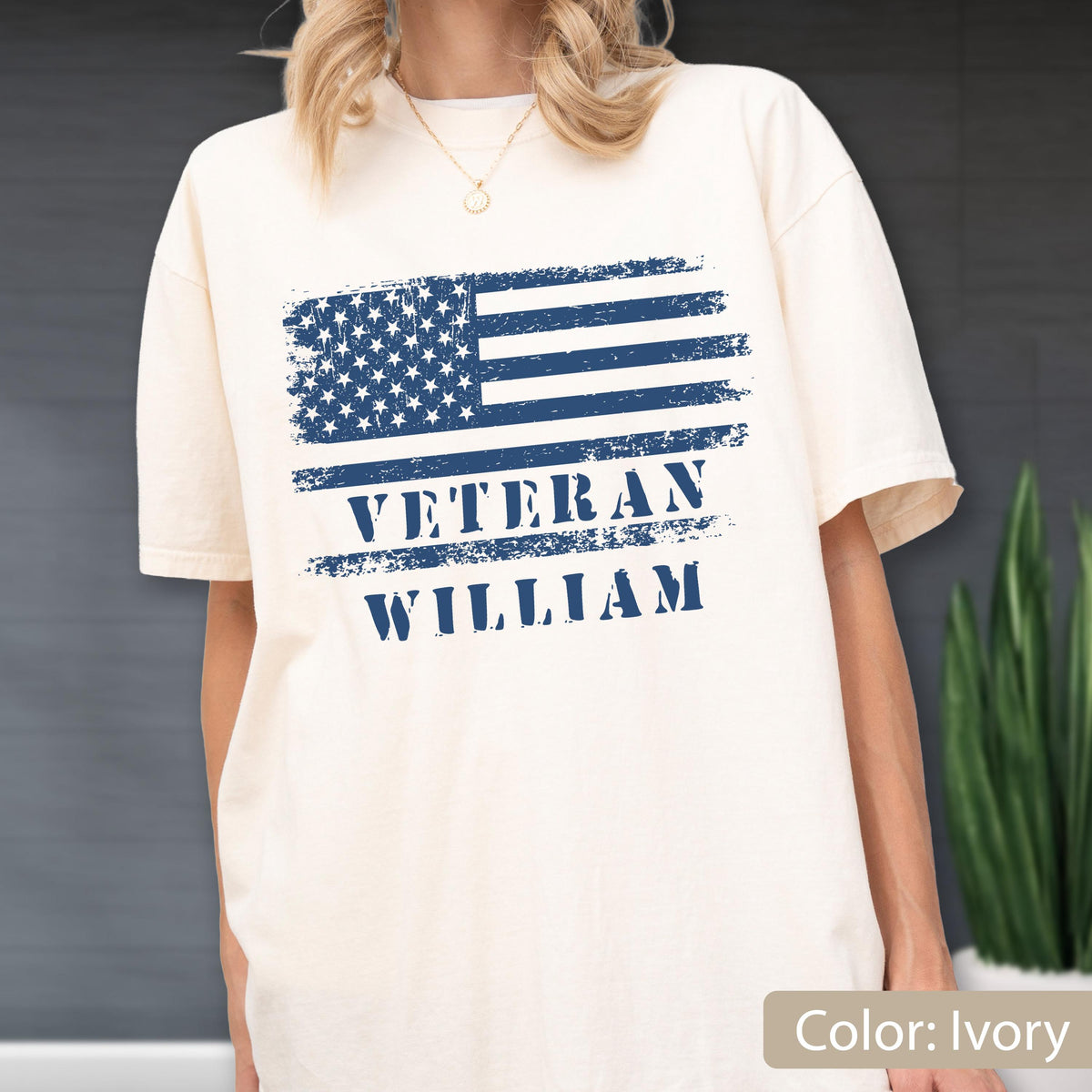 May include: An ivory short-sleeve t-shirt featuring a distressed navy blue graphic of the American flag. Below the flag, the text VETERAN and WILLIAM is printed in a bold, distressed navy blue font, centered on the chest of the garment.