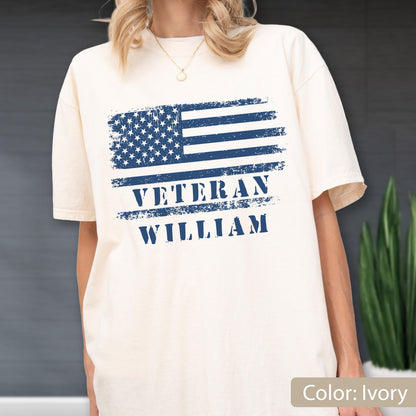 May include: An ivory short-sleeve t-shirt featuring a distressed navy blue graphic of the American flag. Below the flag, the text VETERAN and WILLIAM is printed in a bold, distressed navy blue font, centered on the chest of the garment.