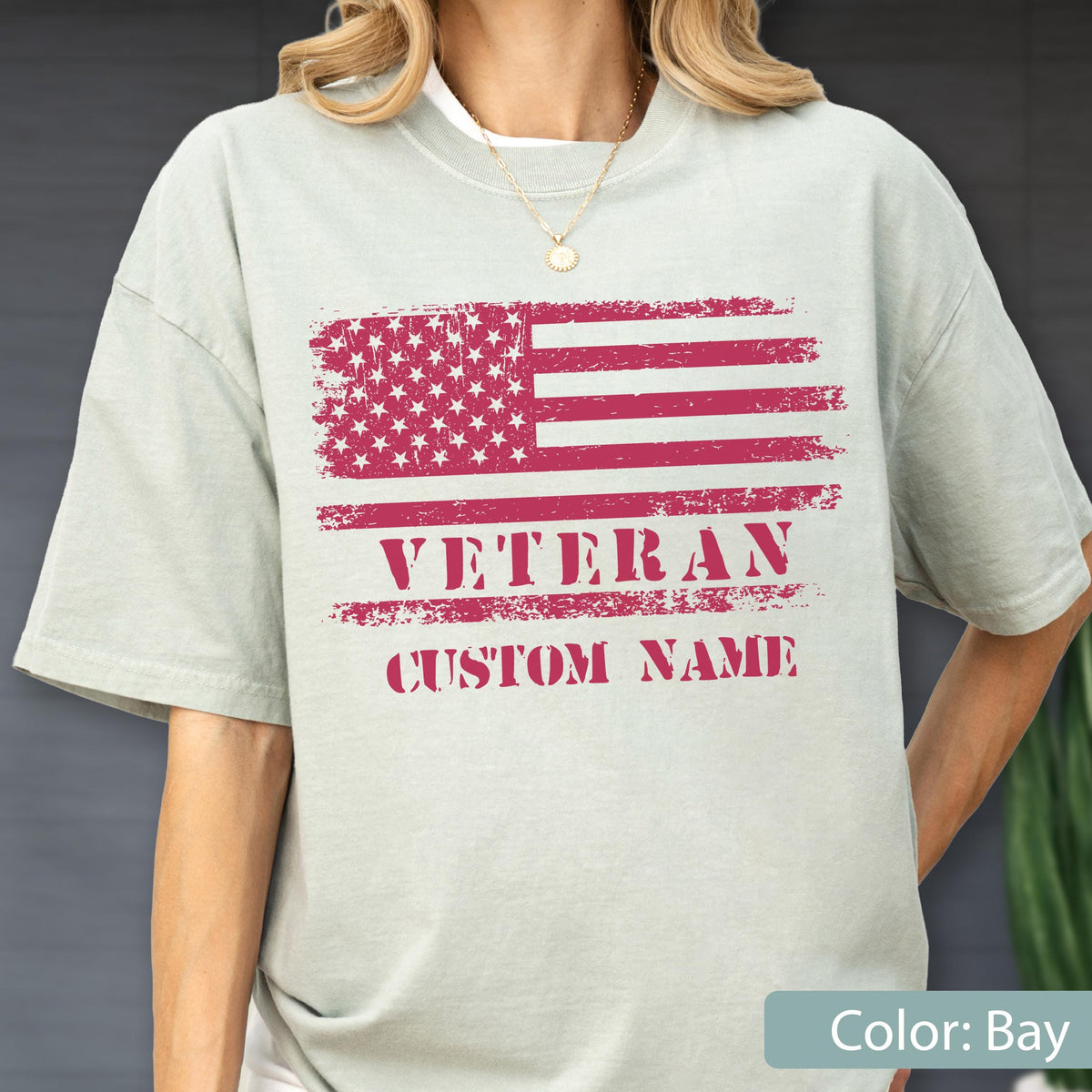 May include: A light sage green short-sleeve t-shirt featuring a distressed red graphic of the American flag. Below the flag, the text reads VETERAN in bold capital letters, followed by the text CUSTOM NAME in a matching red distressed font.