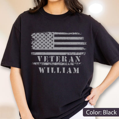 May include: A black short-sleeved t-shirt featuring a distressed grey graphic of the American flag. Below the flag, the text VETERAN is printed in a bold, distressed grey font, followed by the name WILLIAM in the same grey lettering.